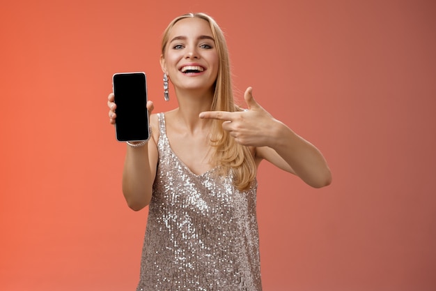 Boastful proud charming elegant blond woman in stylish evening dress show smartphone display proudly pointing mobile phone screen smiling showing photo boyfriend, standing red background.