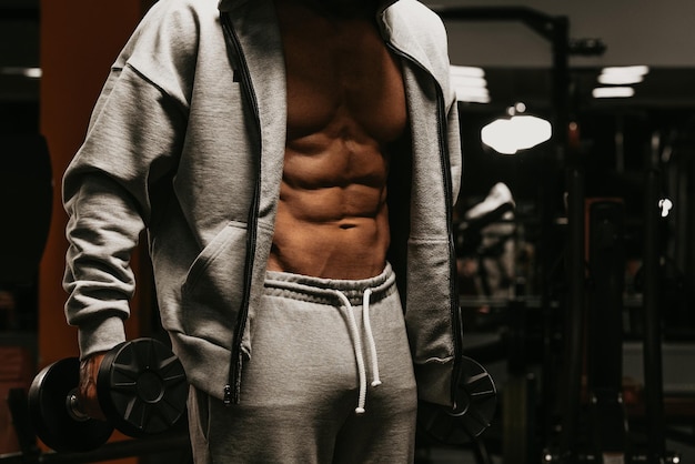 A bodybuilder in his opened zipped hoodie is showing his abdominal muscles while holding dumbbells. A photo of a torso of a sporty guy who is posing after a workout in a gym.