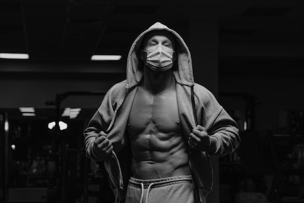 Bodybuilder in a hood and a face mask is opening a hoodie to demonstrate his abs