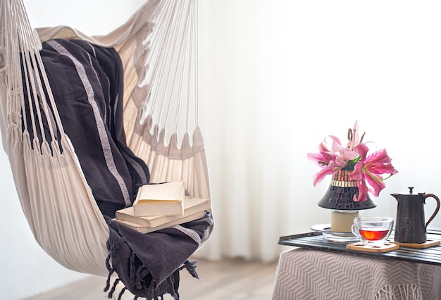 A boho-style hammock chair with a stack of books. Wooden tray with teapot and Cup of tea and beautiful Lily flowers. Cozy interior. The concept of rest and home comfort. Space for text.