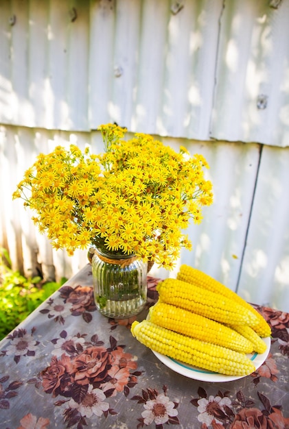 Boiled corn lies on a plate on the table along with a bouquet of yellow flowers Sunny summer day