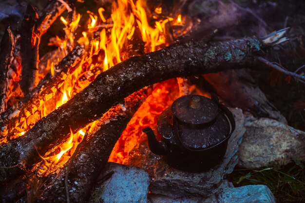 Boiling of tea in kettle on bonfire with large firewood. Tea drinking in open air. Active outdoor recreation. Camping in dusk. Romantic warm atmosphere in twilight on nature. Active rest.