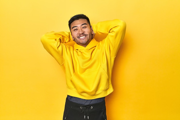 Bold Asian man in yellow hoodie on yellow studio stretching arms relaxed position