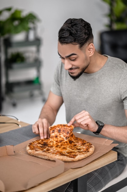 Bon appetit. Attractive dark haired man taking slice of pizza sitting at table at home in good mood