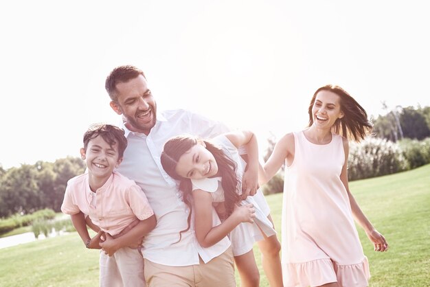 Bonding. Family of four walking on grassy field father carrying kids laughing playful