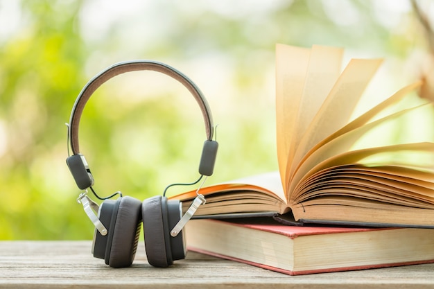 Book and black headphone on wooden table with abstract green nature blur. Reading and education concept