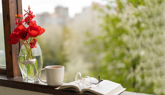 Book, glasses, cup of tea and red tulips on a wooden window