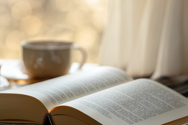 A book is open to a cup of coffee on a table next to a window.