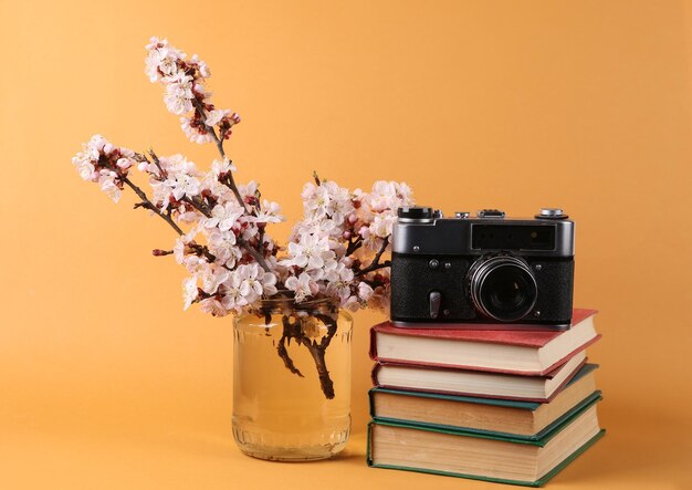 Books and retro camera with Beautiful flowering branches in glass jar on orange background