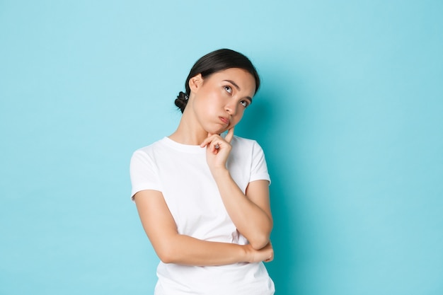 Bored or annoyed cute asian girl in white t-shirt roll eyes, lean on palm and looking indifferent, being not in mood, feeling boredom and apathy, standing blue background not concerned.