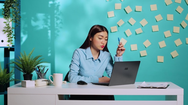 Bored employee working dull monotonous lacklustre tedious office job. Asian businesswoman playing with pen while working on laptop in dreary uninspiring workplace over blue studio background