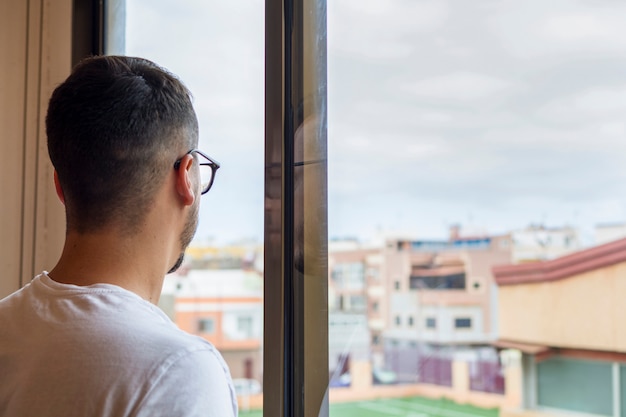 Bored man in quarantine, homebound, looking out the window at the street, from where he sees an empty football ground