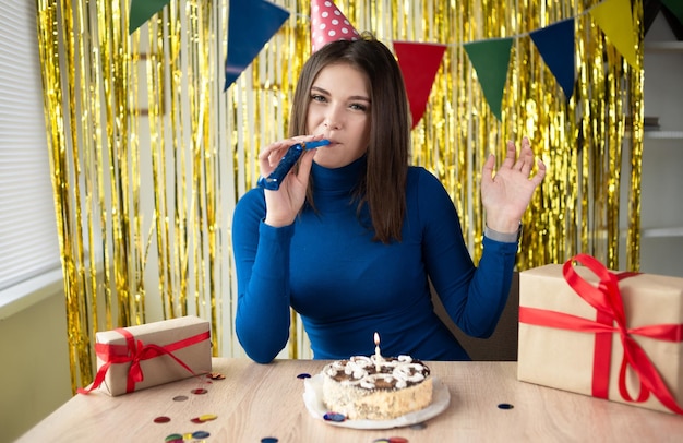 A bored millennial sitting at a table with a cake and gifts blowing a tune in the room against the backdrop of decorations on her birthday is having fun alone A girl in a festive hat at a one party