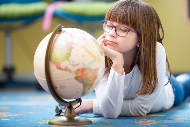 Bored pre-teen girl wearing glasses looking at a globe lying on the floor.