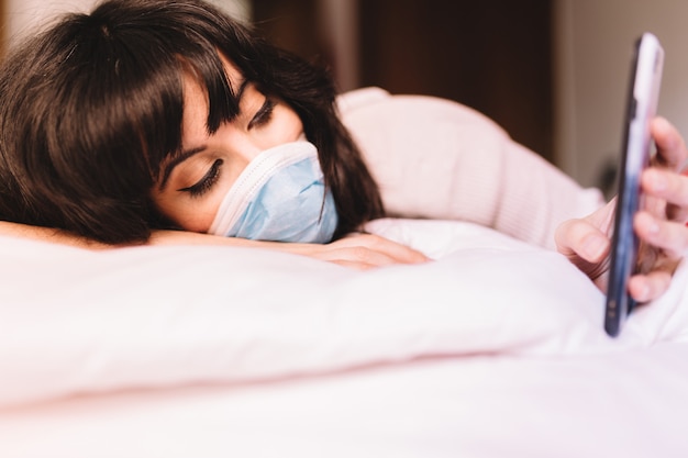 Bored woman at home in breathing medical respiratory mask on her face using mobile phone. pandemic coronavirus, virus covid-19. Quarantine, prevent infection concept. Focus on mobile phone.