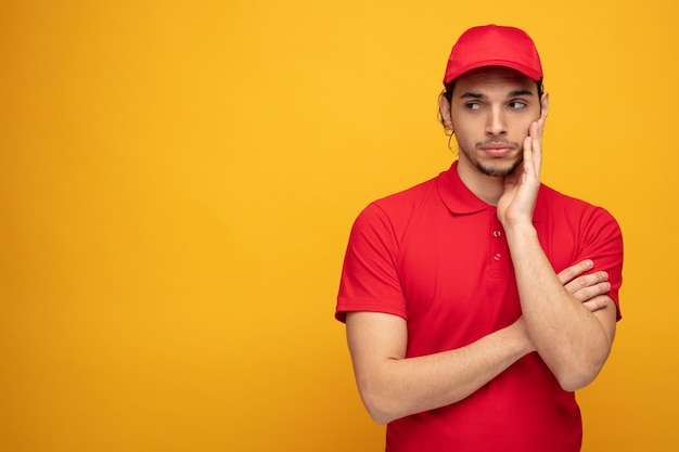 bored young delivery man wearing uniform and cap keeping hand on face looking at side isolated on yellow background with copy space