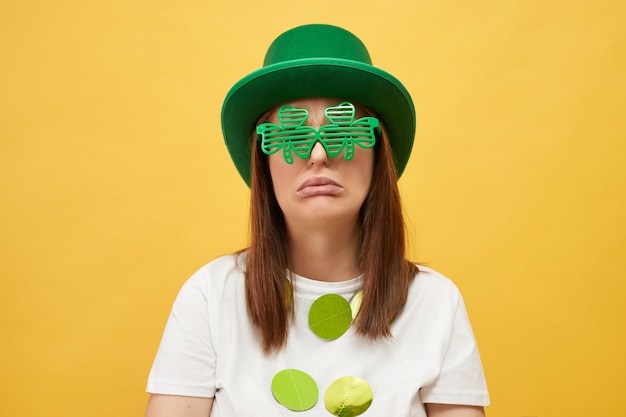 Boring leprechaunthemed holiday party Crying unhappy woman wearing green leprechaun hat and shamrock glasses posing isolated on yellow background looking at camera with pout lips expressing sadness