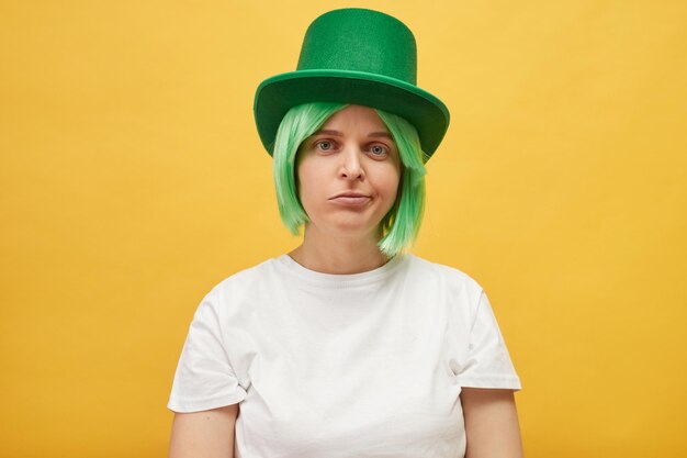 Boring St Patricks Day celebration Sad unhappy upset woman wearing festive green hat standing isolated over yellow background looking at camera with displeased expression