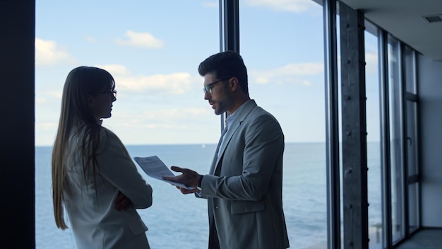 Boss discussing marketing report with stressed businesswoman at panorama window