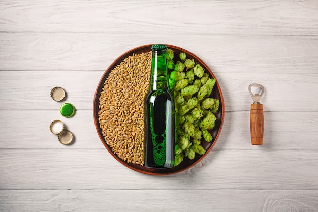 A bottle of beer on a plate with green hops and oat grain with opener and corcks on a white wooden board