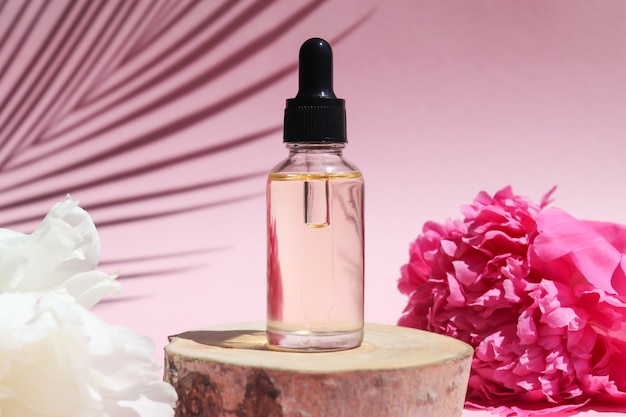 Bottle of cosmetic essential oil with dropper on a wood cut with peony flowers and palm leaf shadow