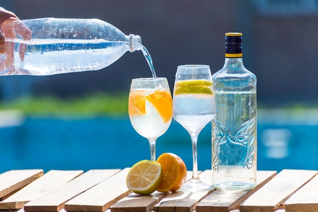 A bottle of gin being poured into two glasses next to a pool