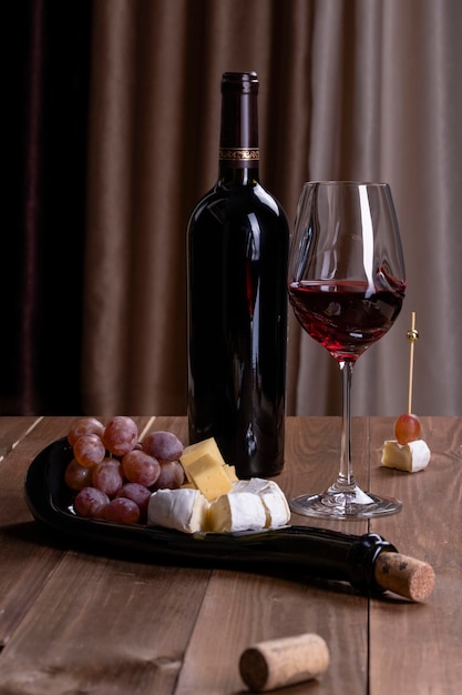 A bottle of red wine a glass with red wine a board with snacks charcuterie board cheese and grapes on a wooden table against a background of brown textile