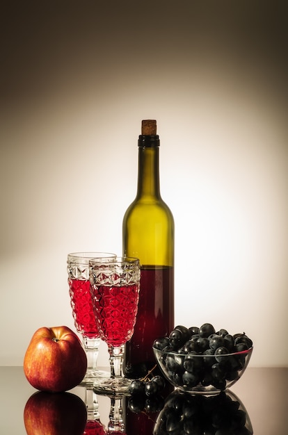 A bottle of red wine two glasses an apple and a cup with grapes standing on a table