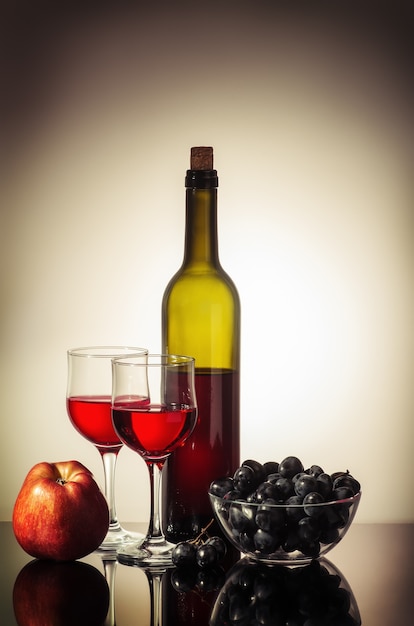 bottle of red wine two glasses an apple and a cup with grapes standing on a table