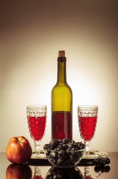 A bottle of red wine two glasses an apple and a cup with grapes standing on a table