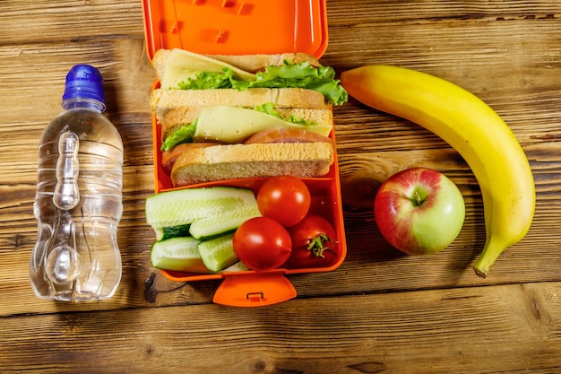 Bottle of water apple banana and lunch box with sandwiches and fresh vegetables on a wooden table Top view