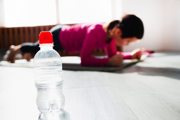 Bottle of water in the foreground. Little girl Doing a Plank Exercise workout at home. Cute kid is training on a mat indoor. Little dark-haired female model has exercises near the window in her room.