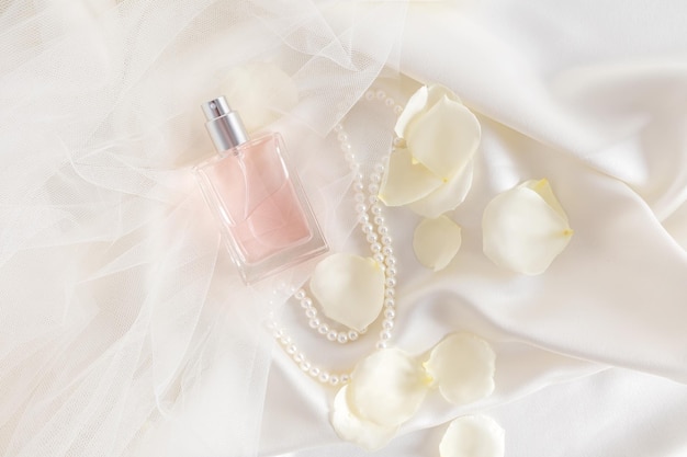 A bottle of women's perfume lies on the bride's wedding veil rose petals and pearl beads satin beige fabric The concept of a wedding fragrance