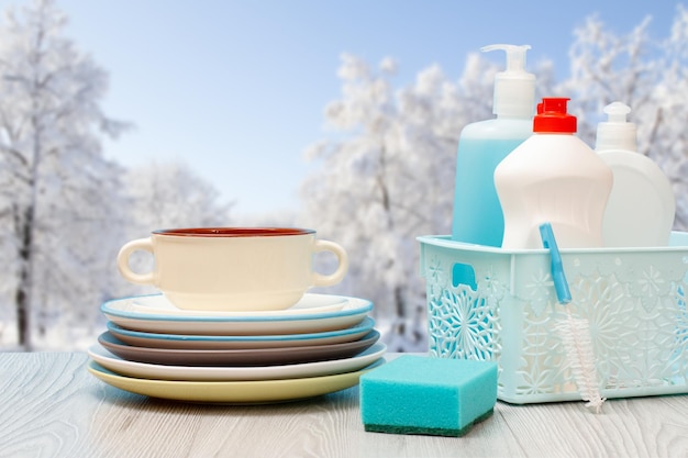 Bottles of dishwashing liquid in basket and plates with winter on background