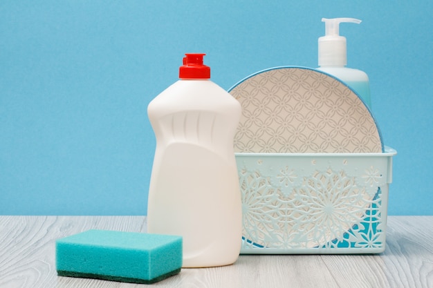 Bottles of dishwashing liquid, plate and sponge on blue background.