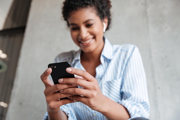 Bottom view of a smiling young african woman wearing shirt listening to music with wireless earphones while sitting at the wall at home and using mobile phone