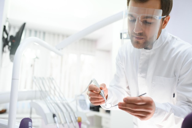 Bottom view young confident happy professional stomatologist european doctor man 20s in white gown hold showing tooth drilling machine at dentist office indoor cabinet Healthcare aesthetic treatment