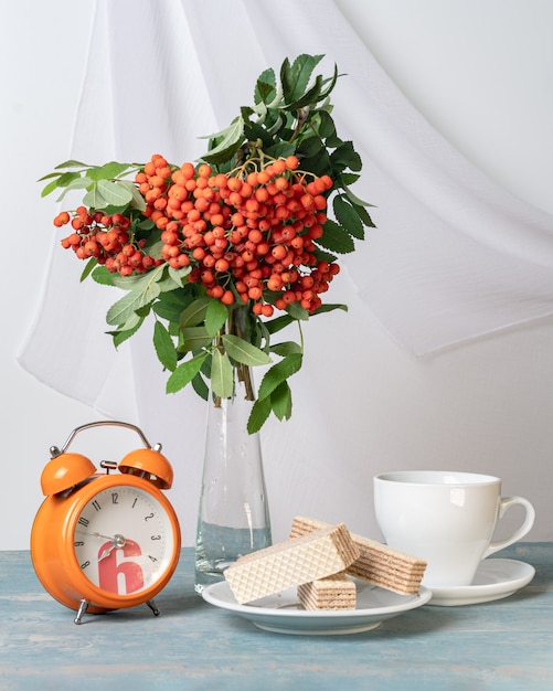 A bouquet of branches with red rowan berries, an alarm clock, a white cup and waffles on a white plate on the table, light key, vertical frame. Morning autumn breakfast. Breakfast for loved ones