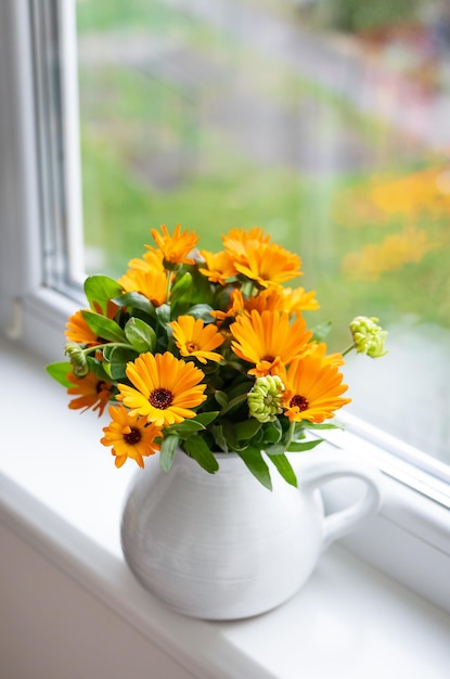 A bouquet of bright calendula in a milk vase stands on the windowsill bright autumn with a window