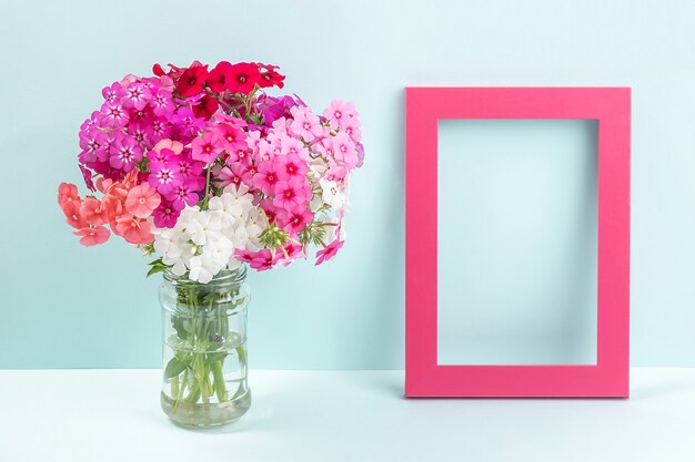 Bouquet of bright colored flowers in vase and wooden empty frame on table against the background of blue wall.