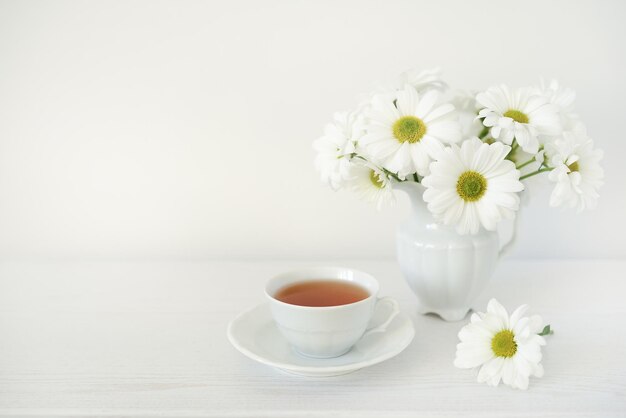 Bouquet of chamomile flowers or daisies flowers and cup with tea on white wooden table, copy space