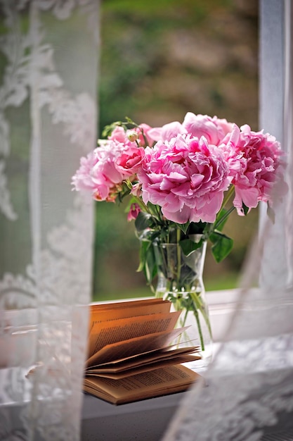 Bouquet of chic pink peonies in a transparent glass vase and an open book on the windowsill