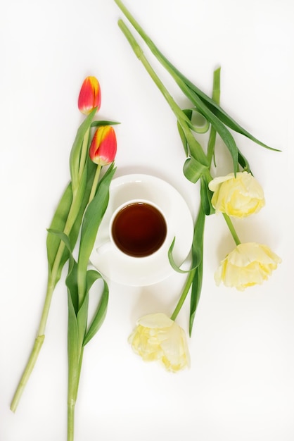 A bouquet of delicate yellow tulips and a cup of tea on a white background Top view The concept of a blog or freelancer Easter Greeting Card