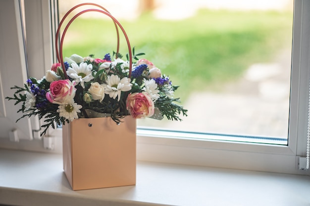 Bouquet of different flowers in paper bag with handles, standing on a windowsill