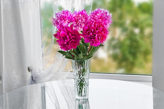 A bouquet of five pink flowers peonies standing in the room on the table in a crystal vase by the window