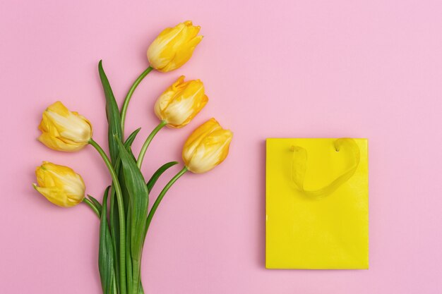 Bouquet fresh flowers of tulip and yellow paper bag on pink backdrop. Spring gift,  holiday background with copy space. Bright colors and minimal style. Top view.