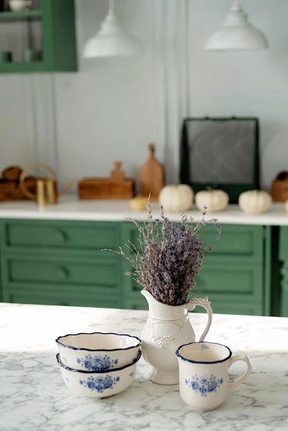 A bouquet of lavender and a set of white dishes a porcelain milk jug a coffee cup and bowls on a white countertop against a green kitchen Soft selective focus