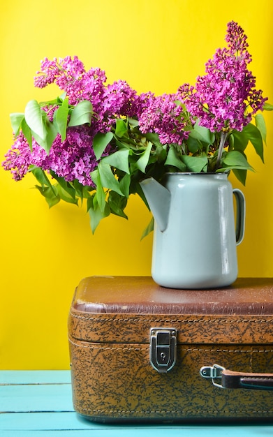 Bouquet of lilac in an old enameled teapot on vintage suitcase on yellow.