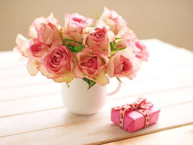 Bouquet of pink roses in white cup on wooden table and gift box