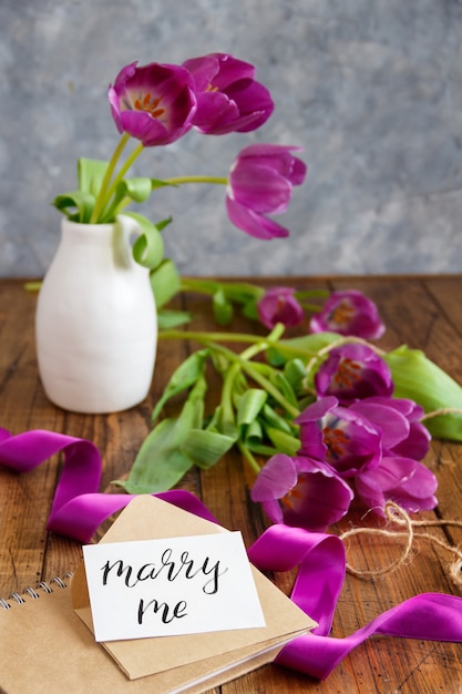 Bouquet of purple tulips and MARRY ME card on wooden table close up